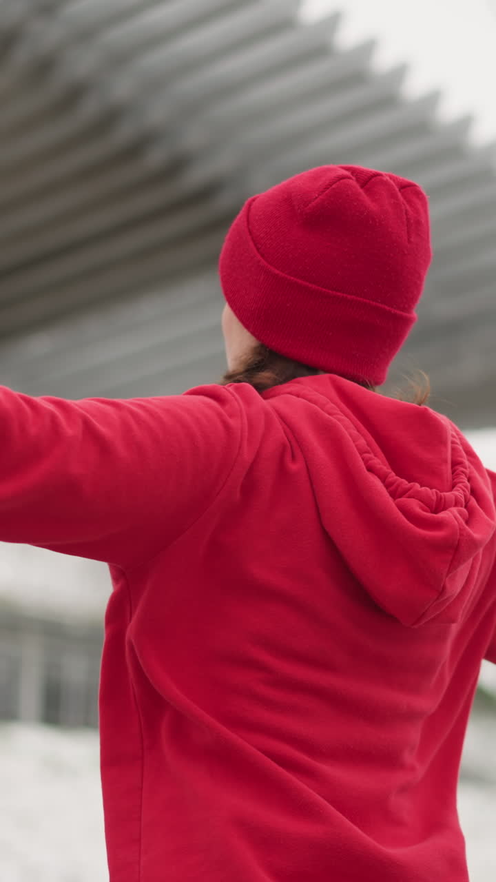 vista trasera de una mujer con guantes estirando los brazos durante el entrenamiento al aire libre bajo un techo de metal con colina nevada y postes de luz en el fondo, mostrando enfoque y energía en un ambiente frío de invierno