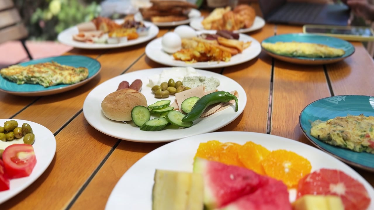 Primo piano di un ricco buffet per la colazione servito su un tavolo da pranzo in legno durante il giorno. Il sano pasto mattutino include cetrioli a fette, olive, uova, salsicce e piatti di frutta fresca.
