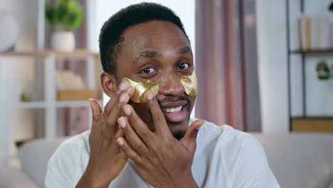 portrait of handsome smiling caring young african american which putting golden patches under eyes to refresh face skin and looking into camera