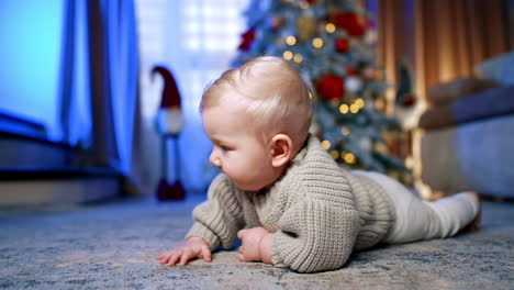 charming caucasian blond infant lies on the floor. little baby boy learning to crawl. christmas tree at backdrop.