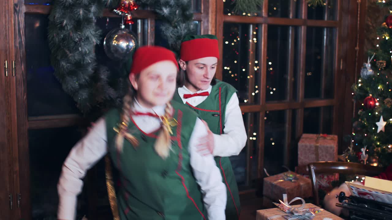 Santa Claus sits at a table with a gift and a typewriter on the background of the fireplace and New Year tree. Dancing christmas elves