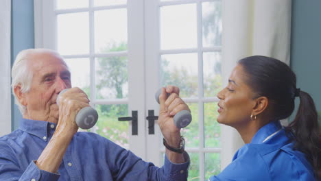 Female-Nurse-Or-Physiotherapist-In-Uniform-Helping-Senior-Man-To-Lift-Hand-Weights-At-Home