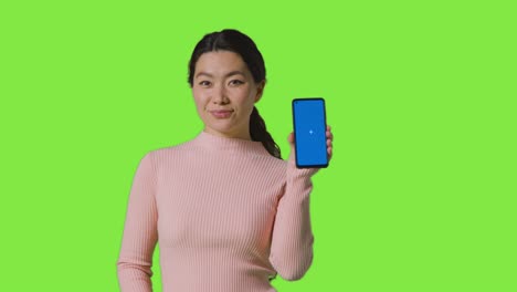 studio portrait of smiling woman holding blue screen mobile phone towards camera against green screen 2