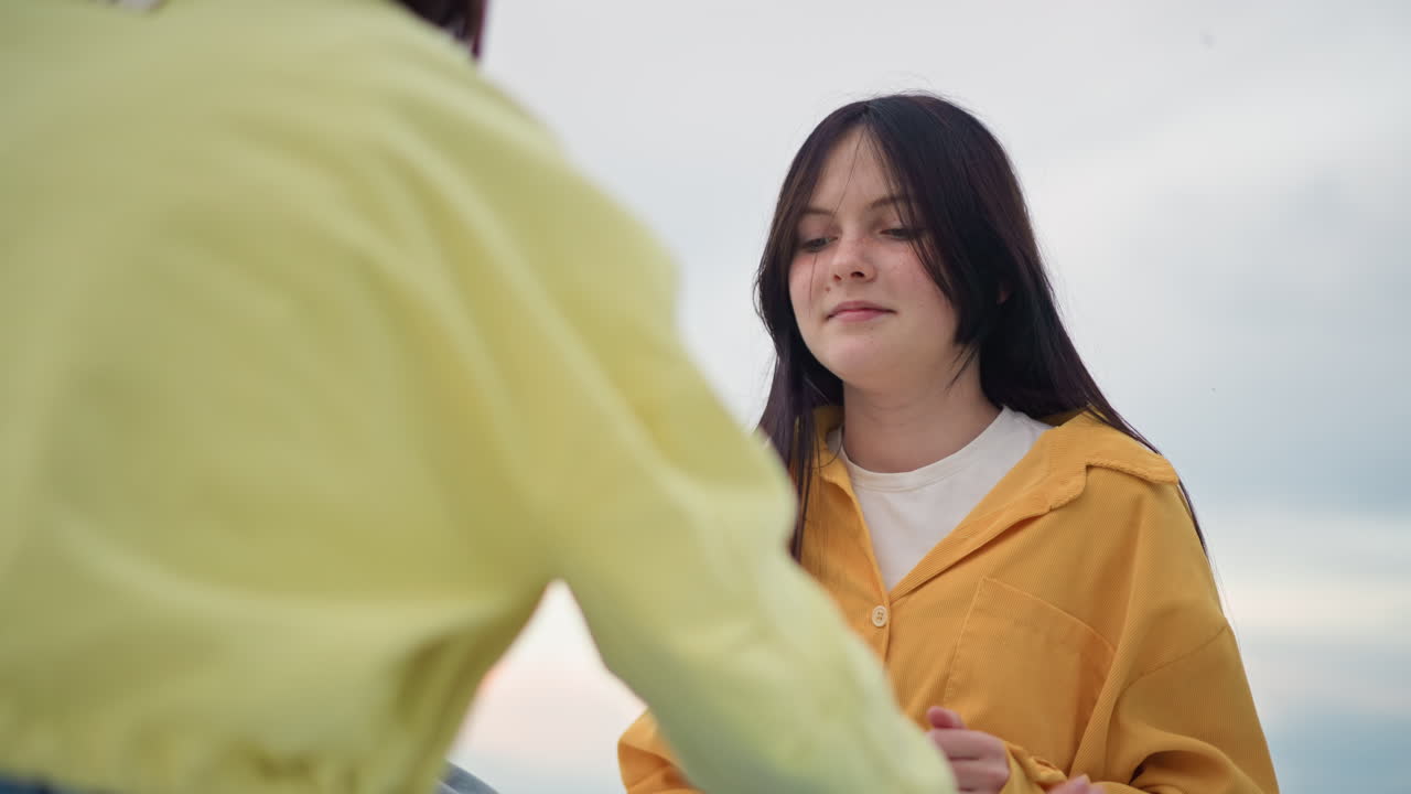 Seorang wanita muda kulit putih mengenakan jaket kuning tersenyum saat bercakap-cakap dengan temannya di tepi laut, rambut dikepang tertiup angin, denim kasual, langit mendung, gestur tangan ekspresif, tawa hangat, gaya hidup apa adanya