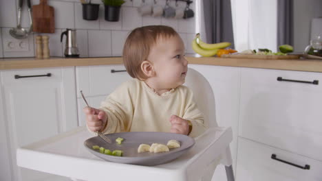 linda niña sosteniendo un tenedor y tratando de comer rebanadas de aguacate mientras se sienta en su silla alta en la cocina