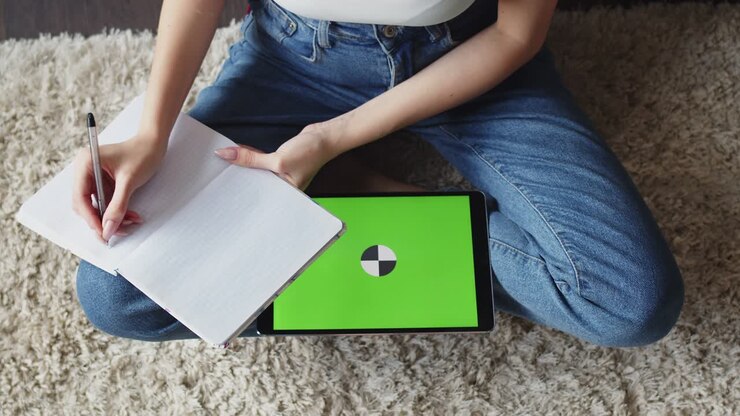 Una donna è seduta per terra e prende appunti su un quaderno, un tablet con schermo verde è appoggiato accanto, vista dall'alto. La donna è seduta e prende appunti guardando lo schermo chroma key del tablet.