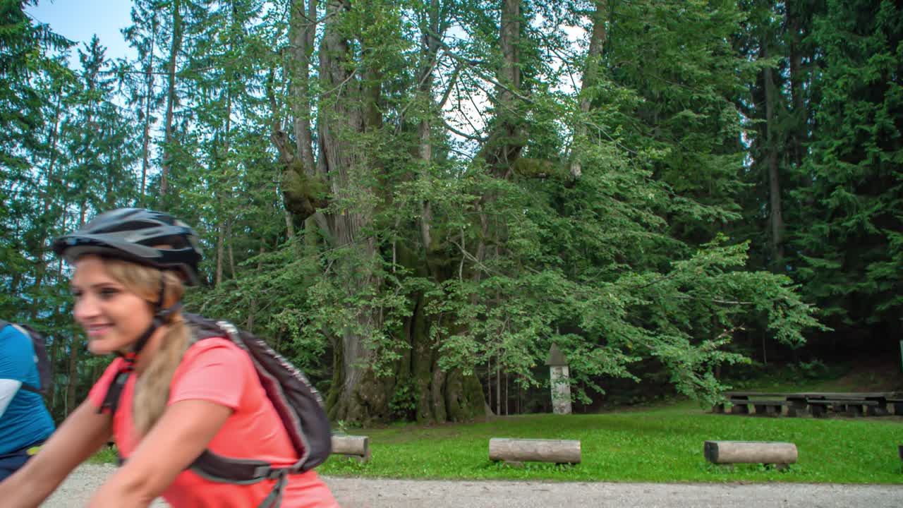 Couple ride bikes on gravel pavement past camera in green forest, slow