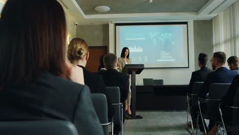 femme d'affaires caucasienne parlant sur un podium dans une salle de conférence et montrant des tableaux et des graphiques sur grand écran