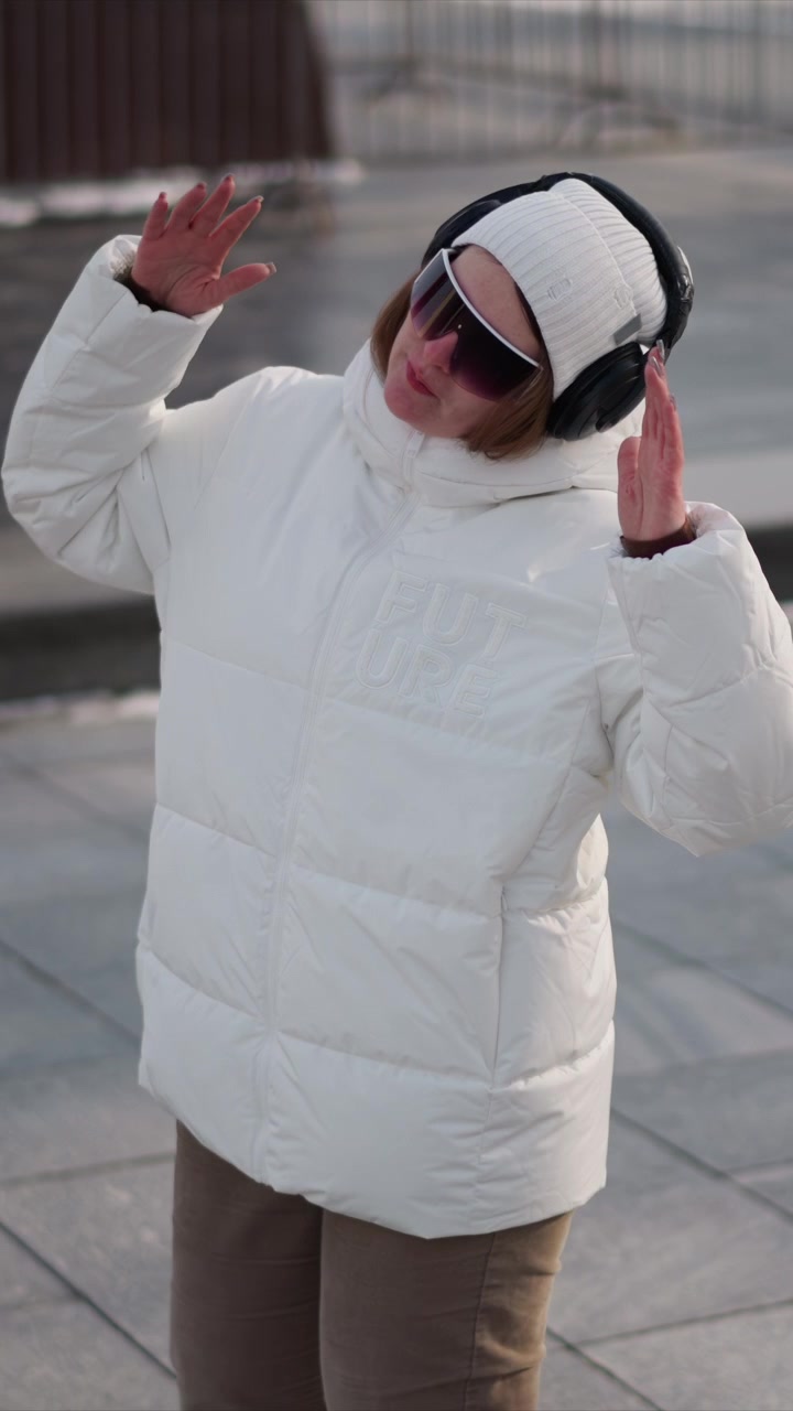 Woman wearing white puffer jacket black headphones and sunglasses dances outdoors on paved ground. Her movements are fluid with arms extended or raised showing expressive hand gestures