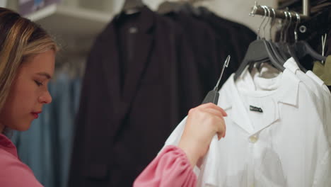 beautiful lady in pink dress retrieves a white shirt from the clothing rack, observes it closely on herself while smiling, shelves filled with neatly arranged clothes are visible in the background