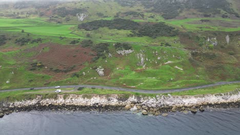 Vista-Panorámica-Aérea-De-La-Idílica-Carretera-De-La-Costa-Rocosa-Justo-En-El-Mar-Con-Coches-En-Movimiento-Durante-Un-Viaje-De-Aventuras-Cerca-De-La-Ciudad-De-Glenarm-En-Irlanda-Del-Norte