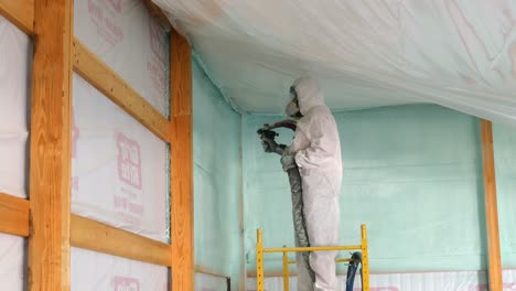 man in disposable tyvek suit and respirator is spraying foam insulation near the ceiling of exterior wall while standing on scaffolding