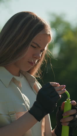 Woman with blonde hair holds green air pump in her gloved hands, inspecting it thoughtfully, against backdrop of lush green trees and natural sunlight