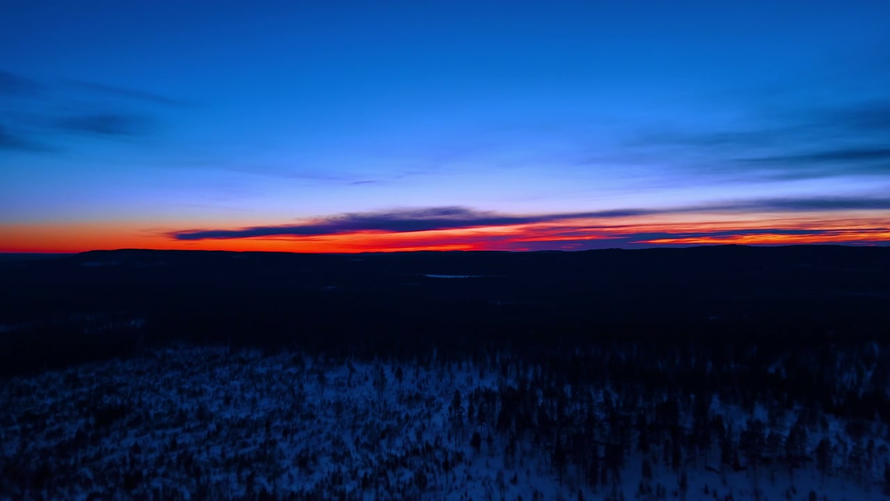 Karla kaplı kuzey ormanı üzerinde alacakaranlık parıltısı, Lapland, Finlandiya. Koyu tayga siluetinin üzerinde kırmızı ve turuncu ufuk şeridiyle derin mavi gökyüzü.