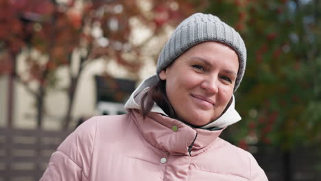 middle-aged woman in pink jacket and grey beanie, smiling gently with head tilted, blurred autumn trees and house blurred in background