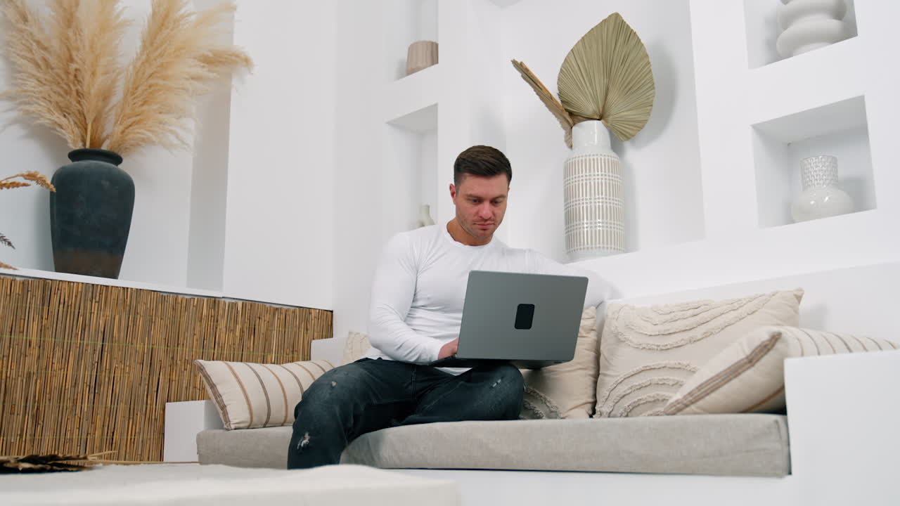 Dark-haired Caucasian male sits on sofa focused on laptop. Working remote from home. Low angle view.