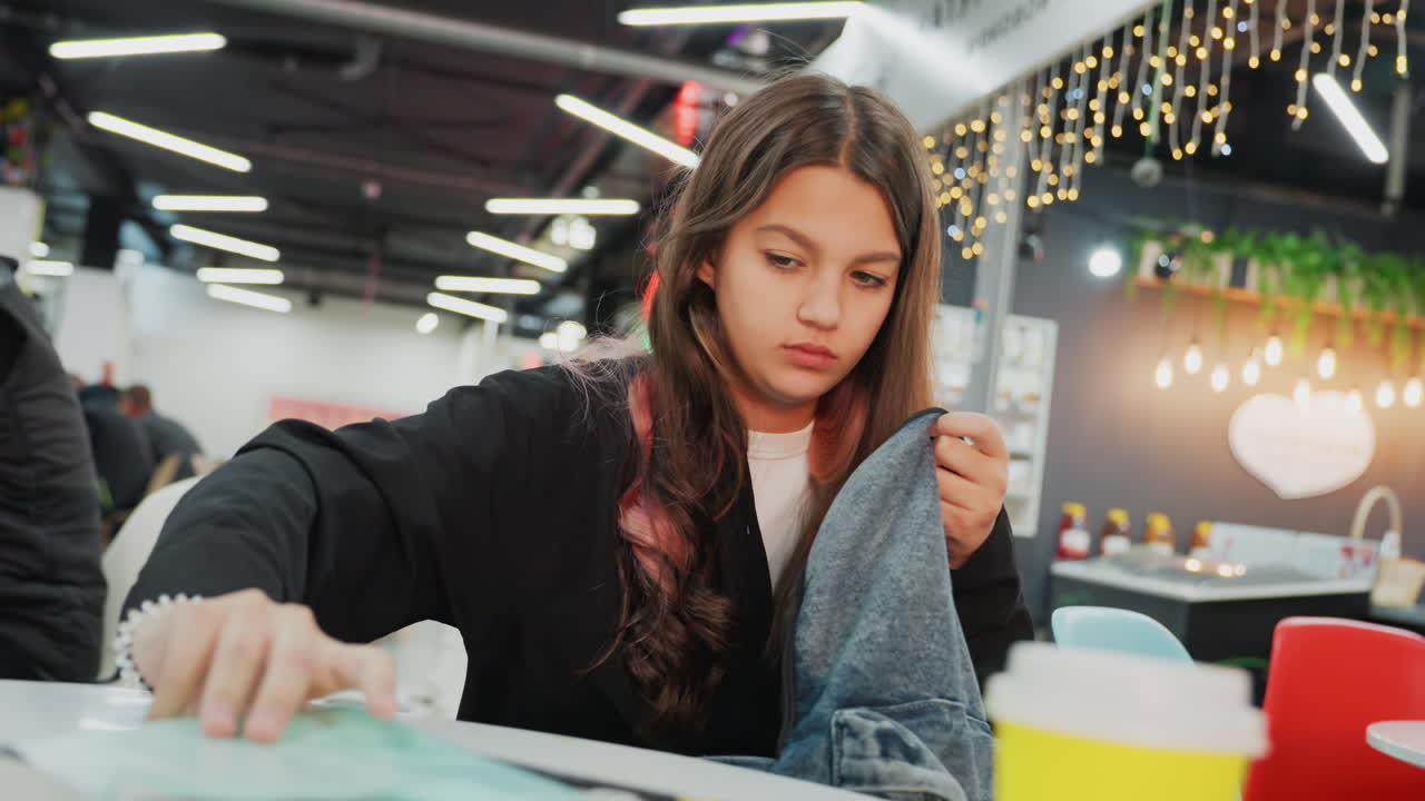 young student studying at cafe table, sorting notebooks, folding sweater, flipping textbook pages by yellow cup, warm string lights and soft bokeh, focused and contemplative preparing for exam