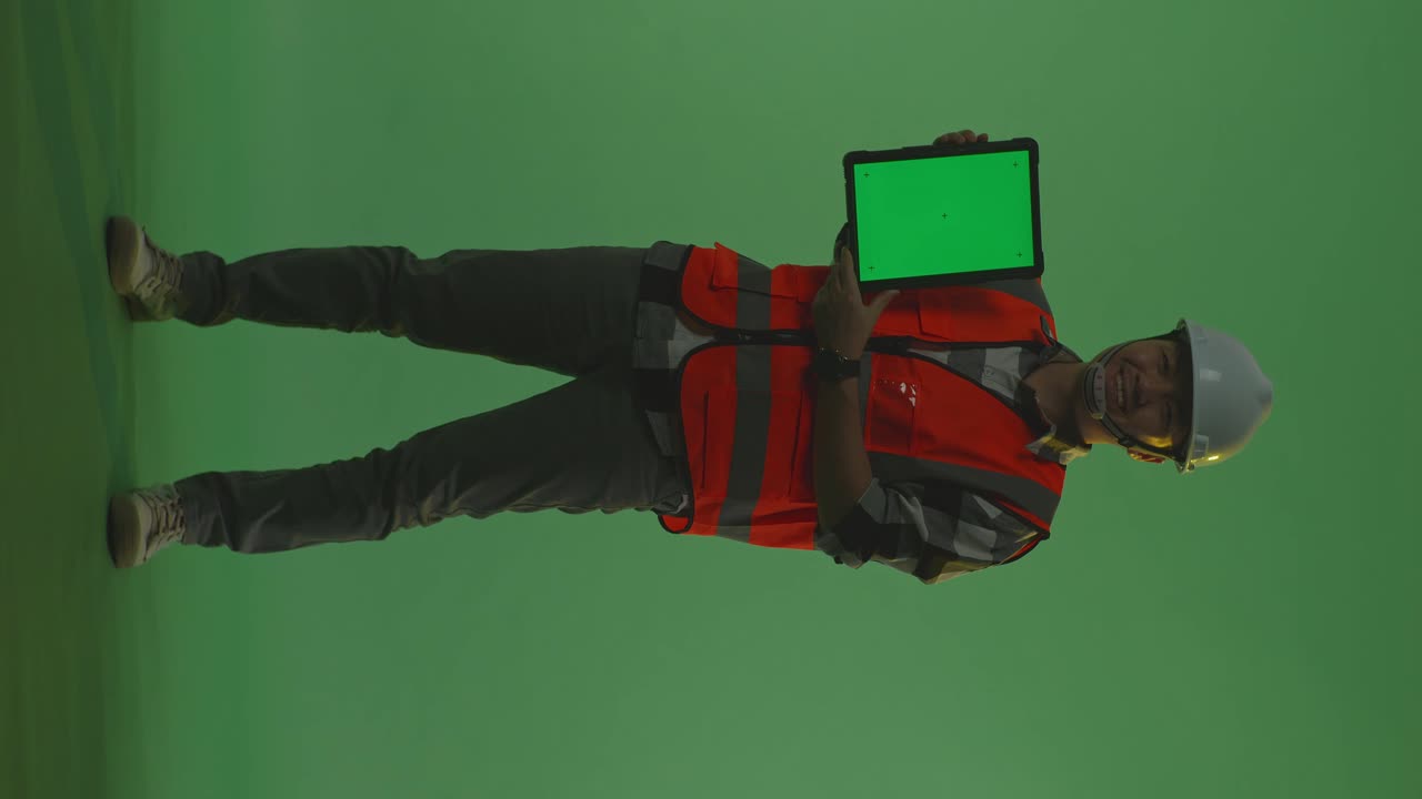 cuerpo lleno de ingeniero masculino asiático con casco de seguridad sonriendo y mostrando la tableta de pantalla verde a la cámara mientras está de pie en el estudio de fondo de pantalla verde, durante la puesta o salida del sol