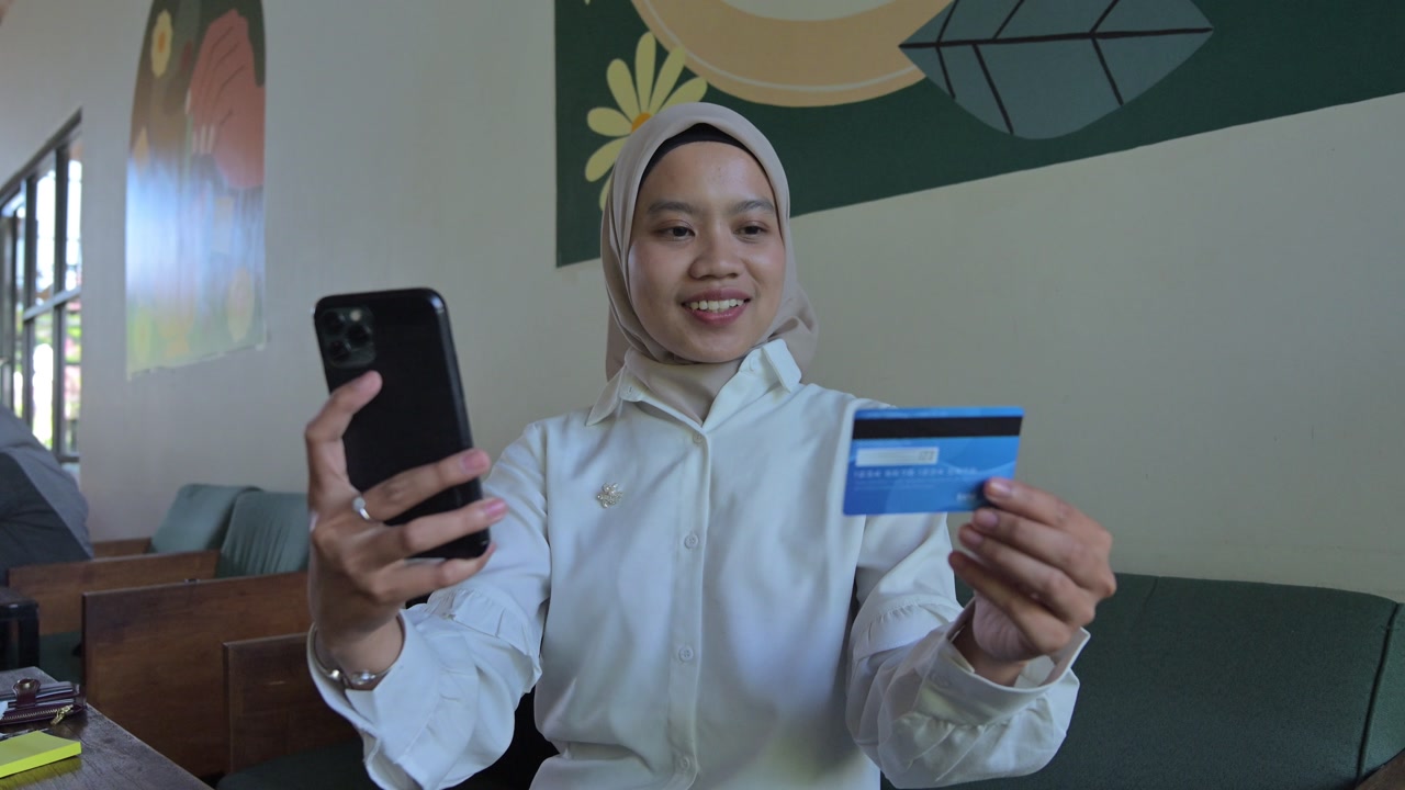 A businesswoman, an Asian woman wearing a hijab, holds a smartphone and a credit card. She appears to be shopping online.