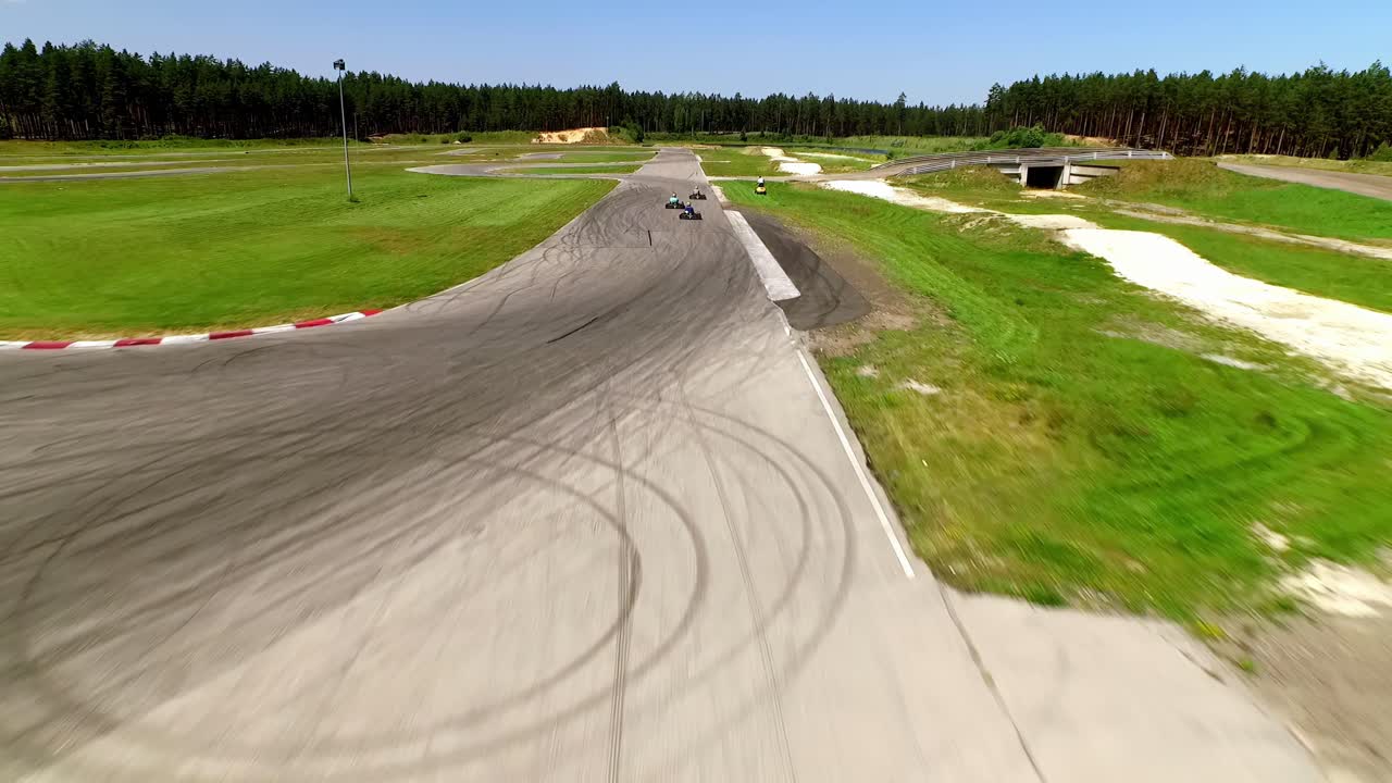 Trase 333, sunny summer day, aerial view with a wide-angle lens, capturing go-karts racing on the gray asphalt track, surrounded by vibrant green grass and a dark forest backdrop