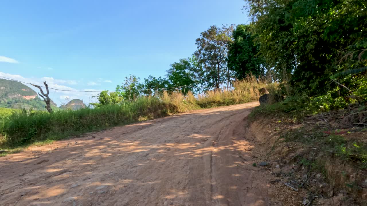 Vehicle travels on winding dirt road through lush, sunlit landscape with distant limestone hills
