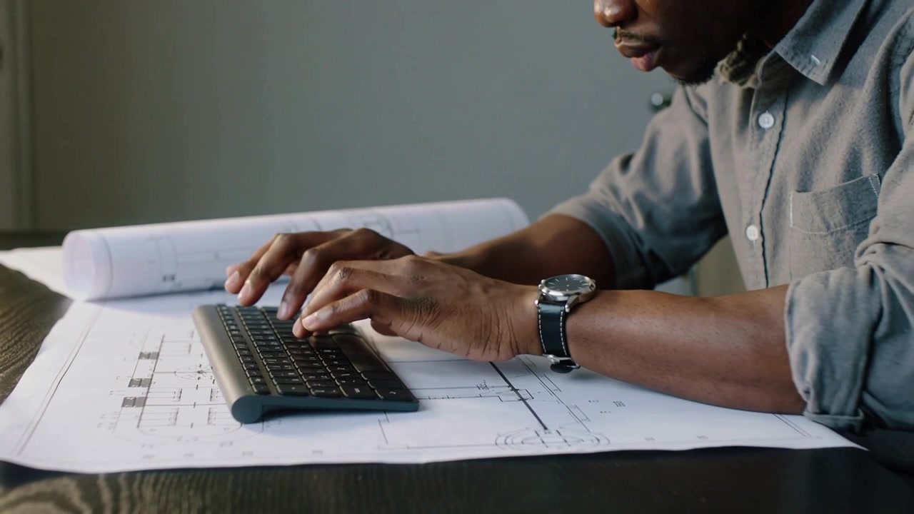 Typing man in rolled sleeves at studio desk updating plans on keyboard, camera revealing hands