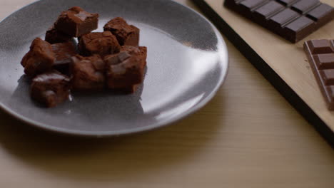 close up of person picking up steaming cup of hot chocolate drink on table next to plate of brownies and chocolate bars