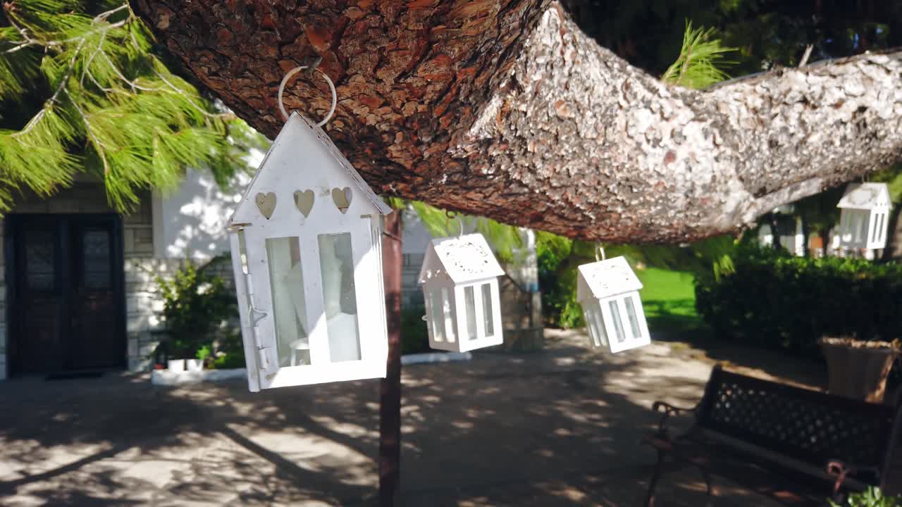 Swaying in the wind cases for candles suspended by a tree branch, garden and house on the background in Nikiti, Greece. Slow motion