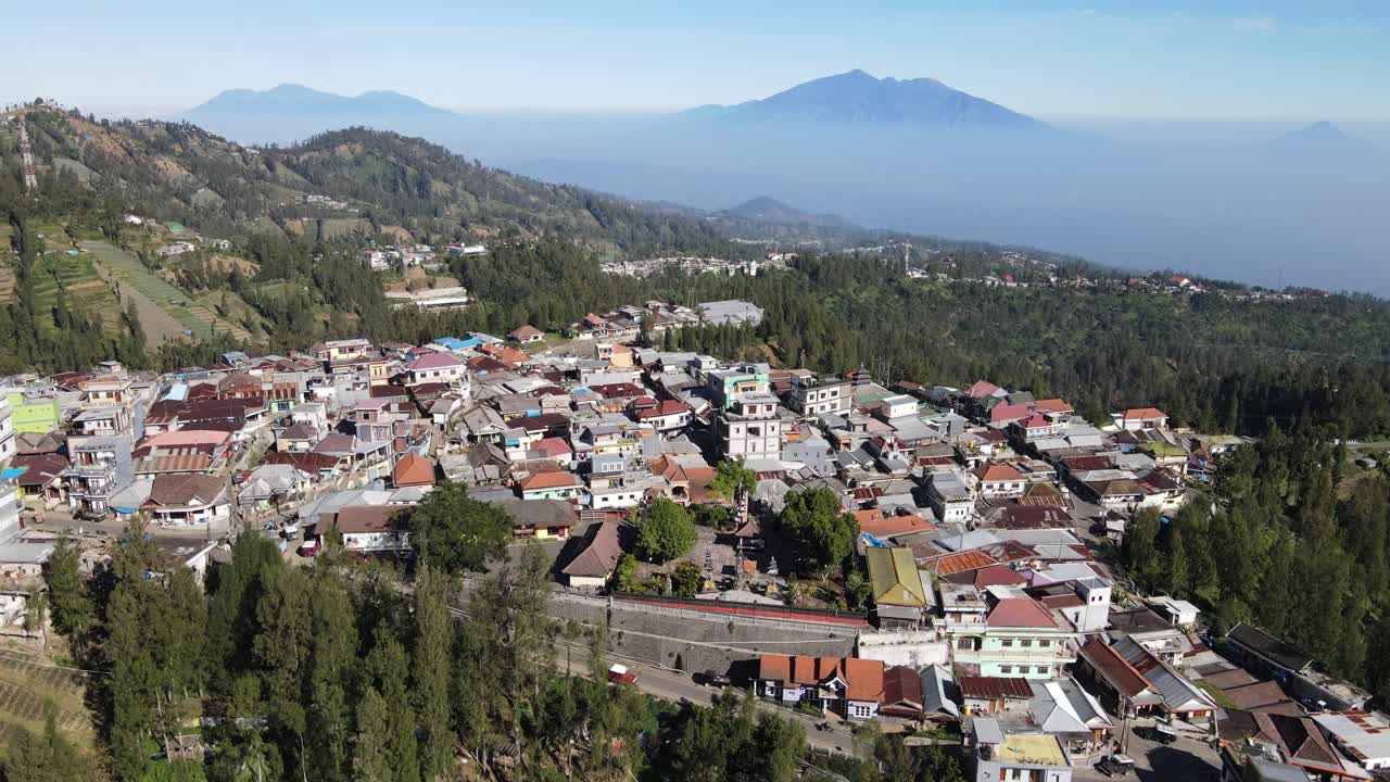 Aerial drone footage of Indonesian mountain town with colorful rooftops, hillside houses, and scenic valley view with distant volcano in background