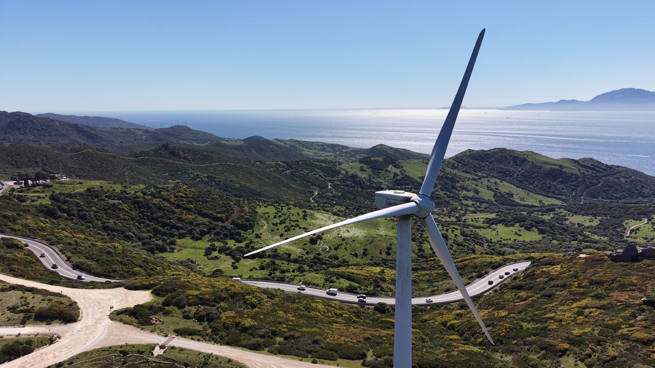 Drone tracking shot of a single wind turbine with a winding mountain road and the African coastline visible across the Strait of Gibraltar