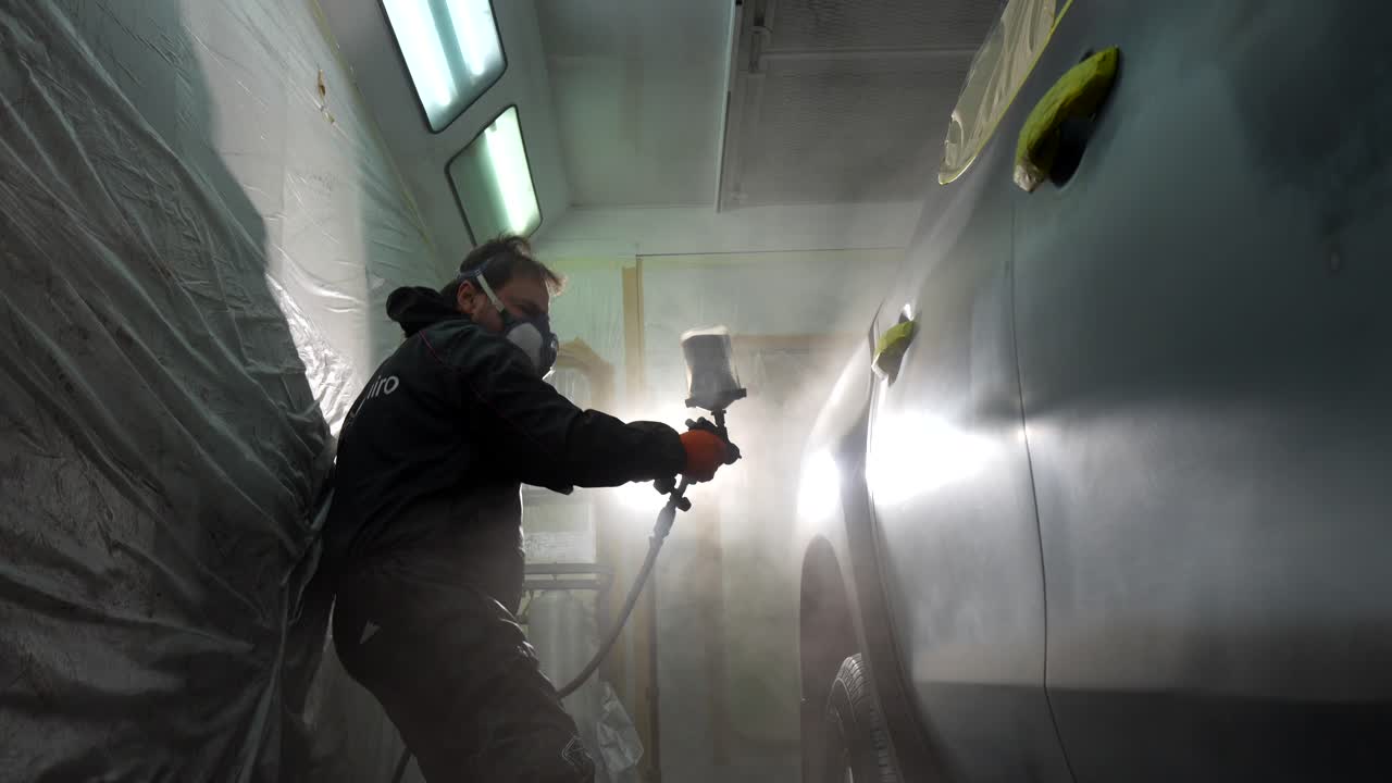 Técnico de carrocería pintando un coche con spray en una cabina de pintura profesional.