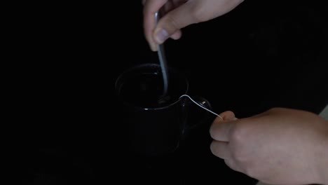 a closeup of a mixed race african american woman's hands and a black tea cup as she holds the tea bag string and gently stirs the steaming hot water as it sits on a stove.