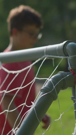 a close-up of a young goalkeeper in red adjusting his glasses after missing a shot that goes into the net, with a blur view of an elderly man in the background