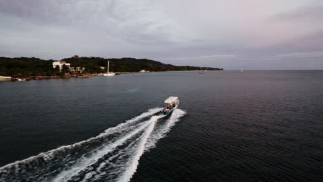 Antena-De-Un-Barco-Motorizado-Deslizándose-Desde-La-Costa-Hacia-Una-Isla-Durante-El-Crepúsculo-En-Roatán,-Honduras