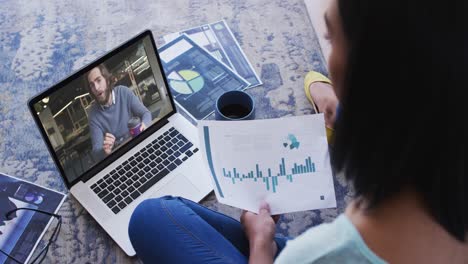 Mixed-race-businesswoman-sitting-on-floor-using-laptop-having-video-call-with-male-colleague