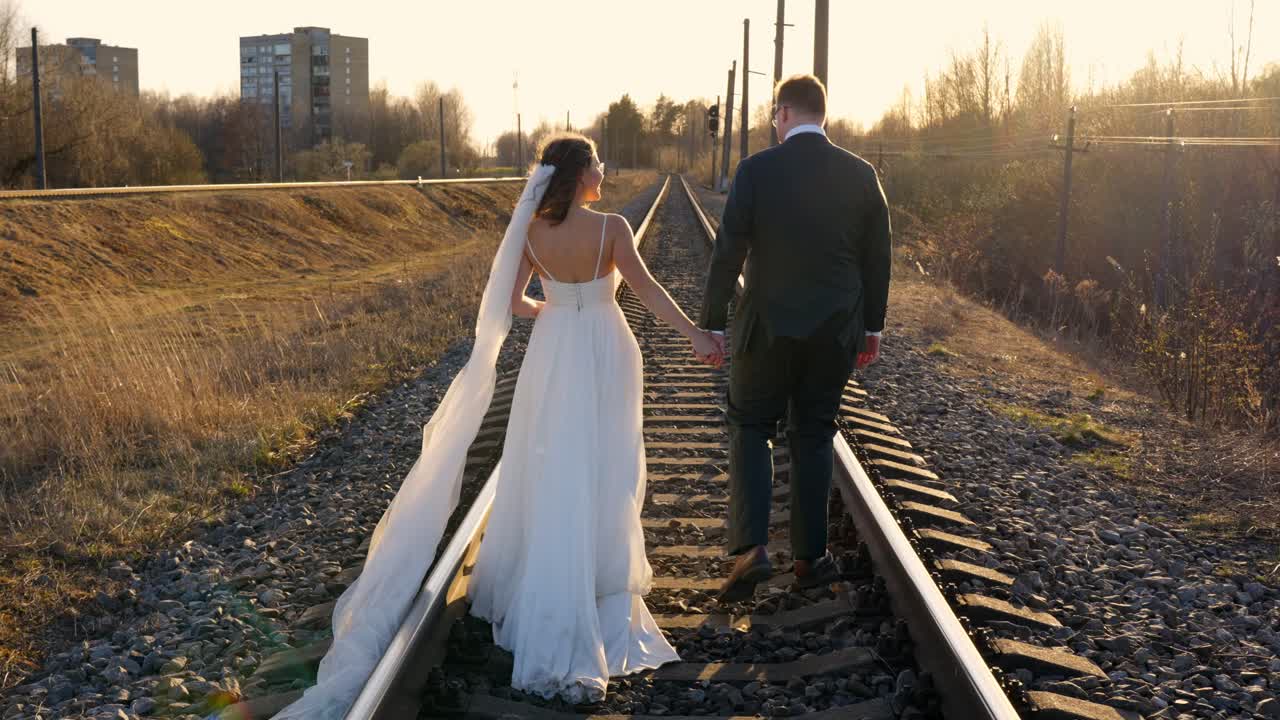 Couple holding hands walking along railway tracks during sunset, wedding theme