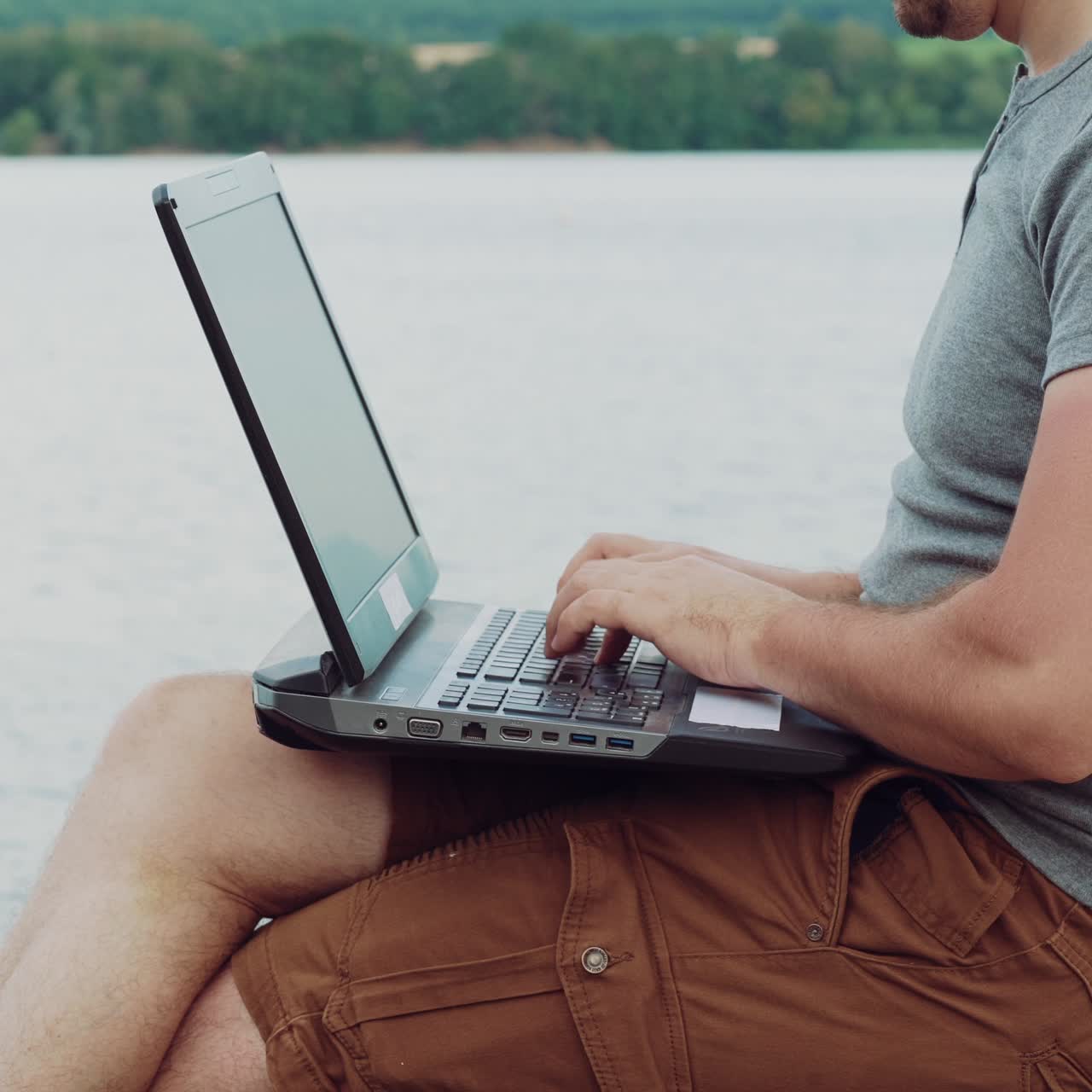 busy man in a gray t-shirt is sitting on a masonry and typing text behind a laptop on the background of nature