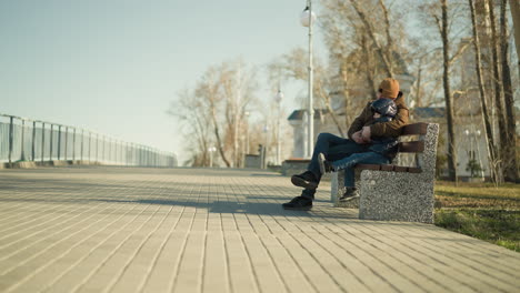 a father and son sit closely on a park bench, the father resting his head on his son's head while holding him with his leg crossed, the son stretches out his left leg