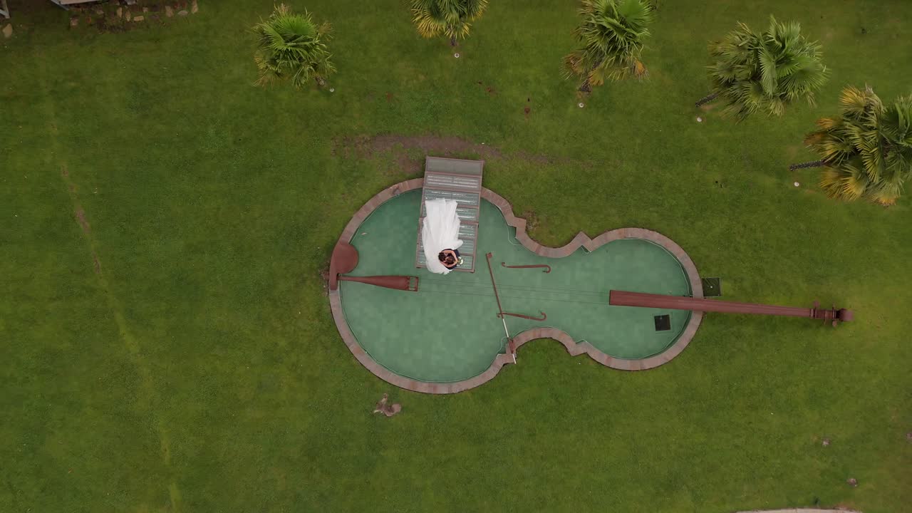 Drone view of a couple standing on a bridge over a violin-shaped lake in an elegant garden.