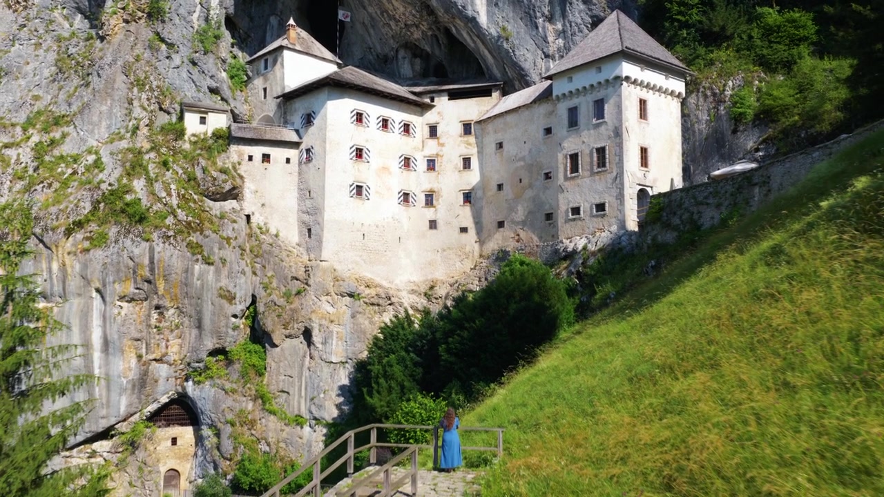 Antico Castello di Predjama con una donna in abito blu in piedi su un punto panoramico in Slovenia. Ripresa aerea in allontanamento.