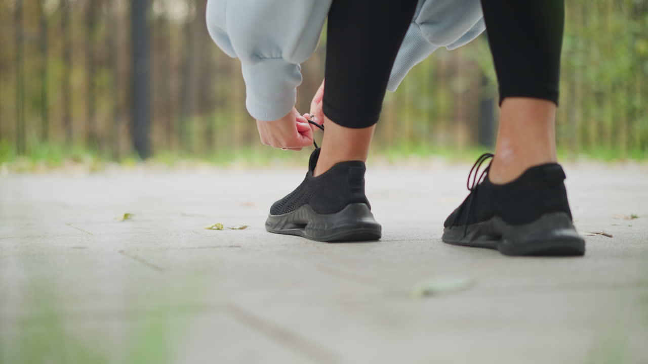 Back view of lady in black leggings and blue sweater tying her left shoe lace on sidewalk in park, preparing for a workout or run outdoors, focusing on active lifestyle and fitness