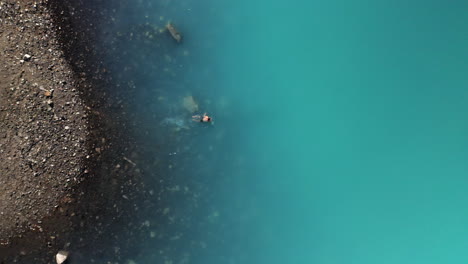 Cinematic-drone-shot-of-man-swimming-in-turquoise-colored-water-in-the-mountains-at-Naltar-Valley-in-Pakistan,-downward-angled-aerial-shot