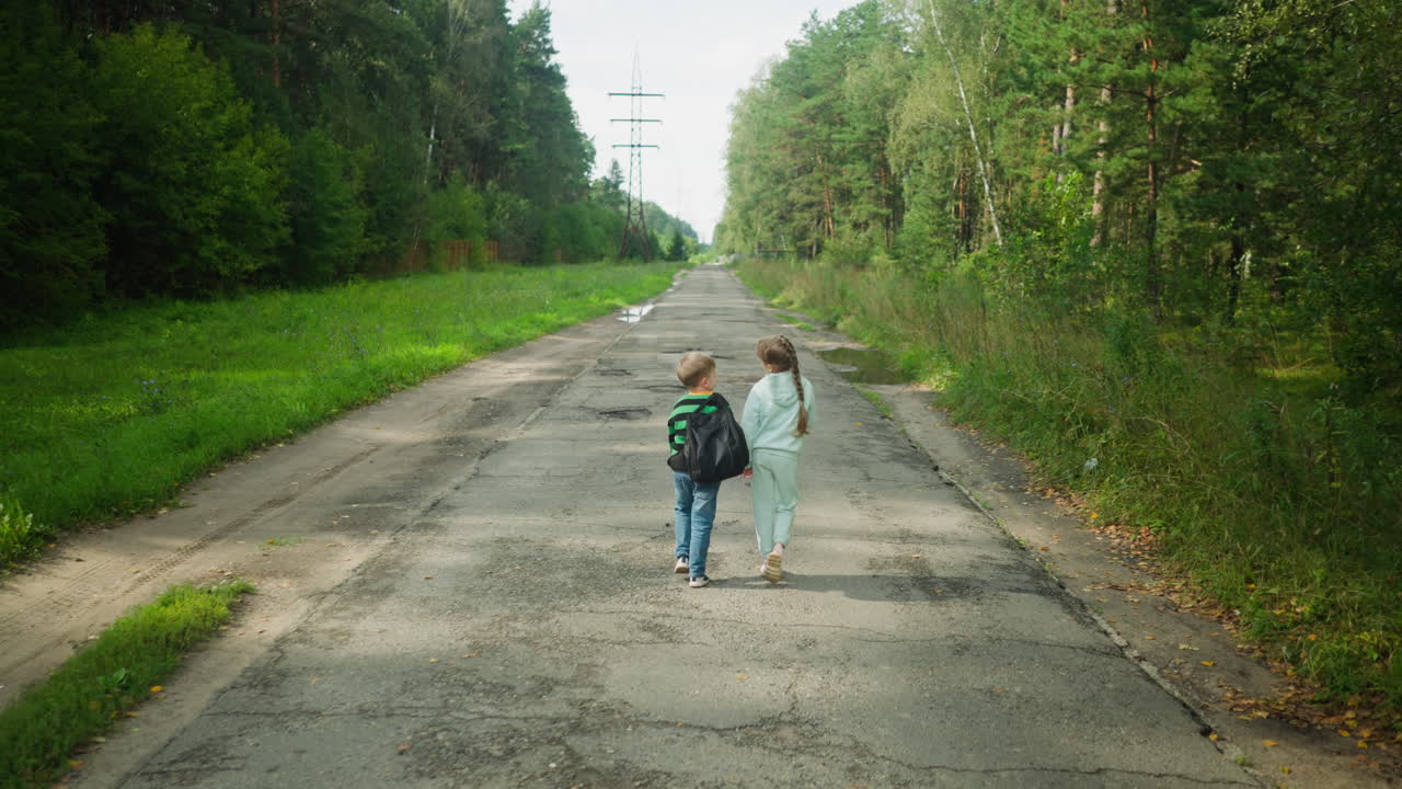 Rear view of siblings walking on cracked paved road surrounded by lush forest, boy engaging in conversation with sister while passing puddle filled potholes under power lines