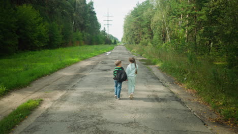rear view of siblings walking on cracked paved road surrounded by lush forest, boy engaging in conversation with sister while passing puddle filled potholes under power lines