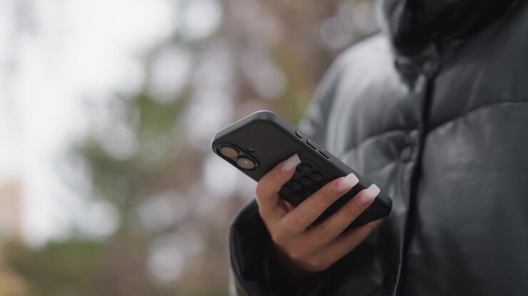 Close up of woman holding and interacting with smartphone in her hand. woman wears black leather jacket showcasing long manicured nails as woman uses device. Soft outdoor lighting creates