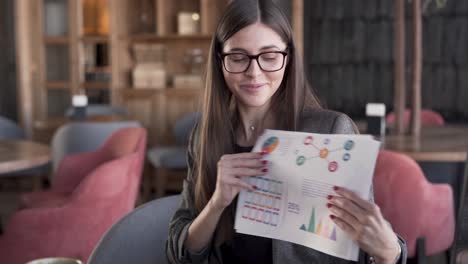 Attractive-young-woman-is-sitting-in-a-cafe-and-holding-a-diagrams-and-statistics-data-in-her-hands