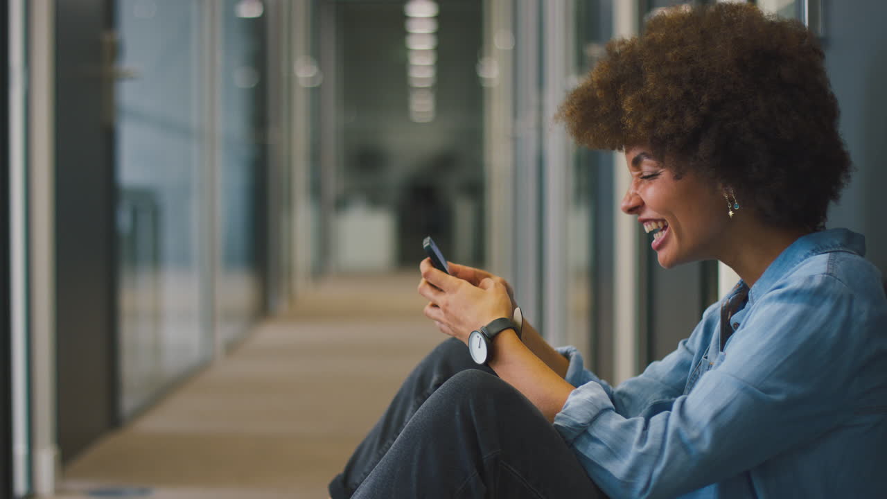 Giovane donna d'affari seduta per terra nel corridoio di un ufficio moderno con il telefono che celebra una buona notizia.