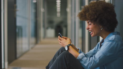 Young-Businesswoman-Sitting-On-Floor-In-Corridor-Of-Modern-Office-With-Phone-Celebrating-Good-News