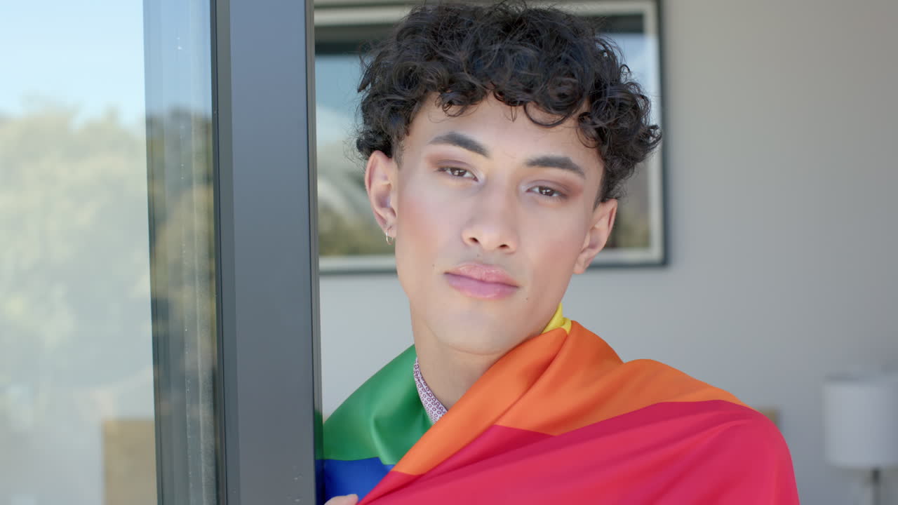 Wearing pride flag, non binary person standing by window, looking confident
