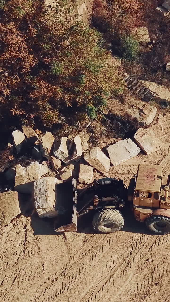 yellow bulldozer with a bucket is carrying a large stone near a sand quarry. Camera motion down. Close-up. Vertical video